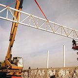Żuraw podnosi białą kratownicę stalową na placu budowy, widoczny pracownik na podnośniku koszowym w tle, błękitne niebo.