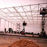 Wznoszenie szkieletu hali z białych profili stalowych, widoczny podnośnik koszowy z pracownikiem na wysokości, w tle krajobraz polny.