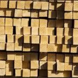 Stos równo ułożonych, suchych, jasnych kantówek drewnianych o kwadratowym przekroju, oświetlonych z góry, tworzących regularny wzór.