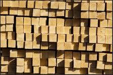 Stos równo ułożonych, suchych, jasnych kantówek drewnianych o kwadratowym przekroju, oświetlonych z góry, tworzących regularny wzór.