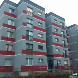 Odnowiony blok mieszkalny z szarą elewacją i czerwonymi pasami, widoczny budynek w trakcie prac pod rusztowaniem i siatką ochronną.