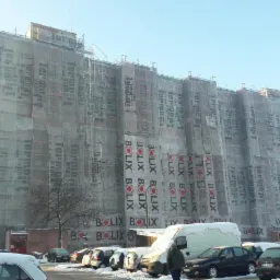 Wielopiętrowy budynek w trakcie prac, osłonięty siatką z nadrukowanymi logo firm Termo Organika i Bolix, widoczny parking z częściowo ośnieżonymi samochodami i dostawczy bus z pokrywą śnieżną...