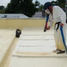 Pracownik w kombinezonie ochronnym natryskuje piankę poliuretanową na dach budynku, widoczne narzędzie natryskowe z długim wężem.