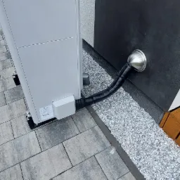 Zewnętrzna jednostka grzewcza Saunier Duval GeniaAir Split HA 12-5 OS 12kW z podłączonym przewodem odprowadzającym skropliny, umieszczona na kostce brukowej obok ściany budynku z zamontowanym...