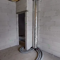 Wnętrze w surowym stanie z widocznymi szarymi, karbowanymi przewodami wentylacyjnymi HeatPex prowadzonymi po podłodze i w górę ściany do rozdzielacza, pionowy kanał owinięty folią aluminiową, sufit...