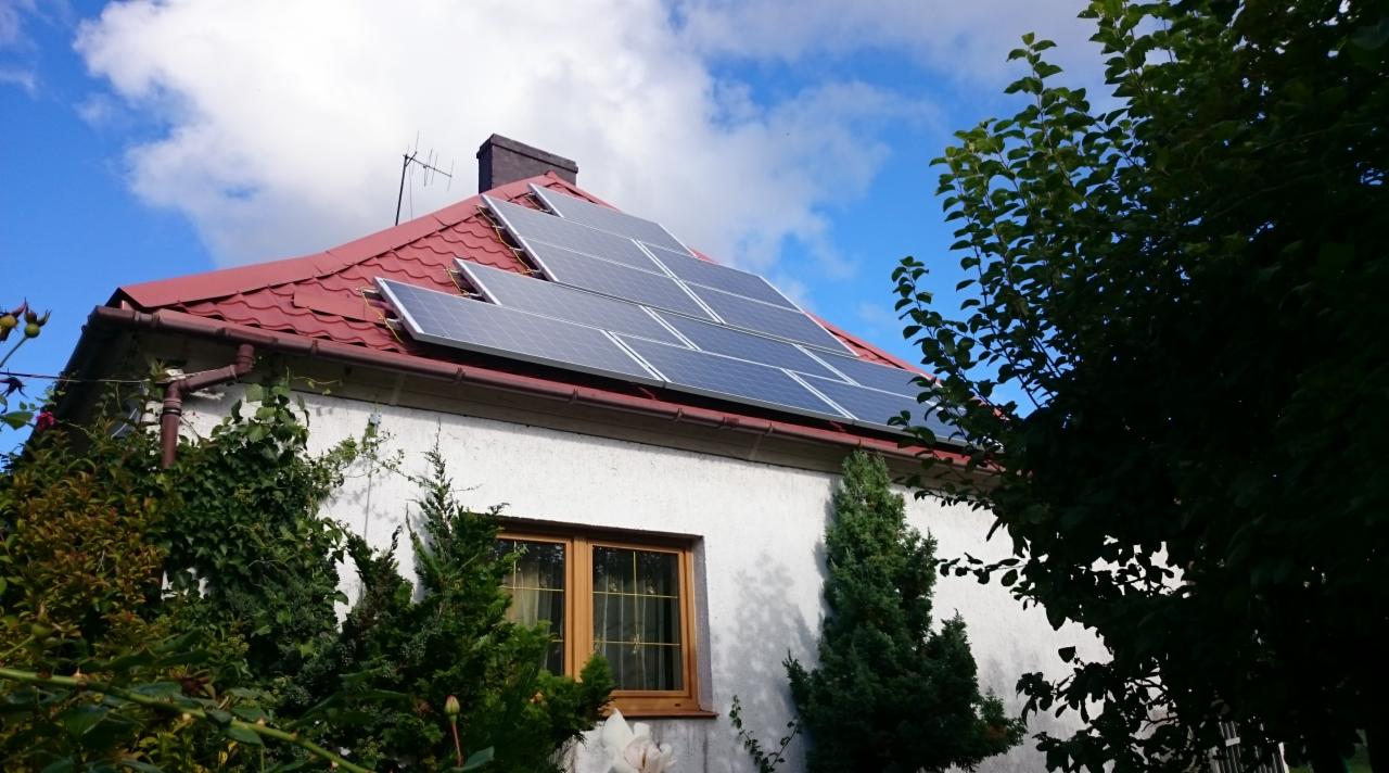 Instalacja fotowoltaiczna na dachu domu jednorodzinnego z czerwoną dachówką, widoczna z poziomu ogrodu z bujną roślinnością, błękitne niebo z chmurami w tle.