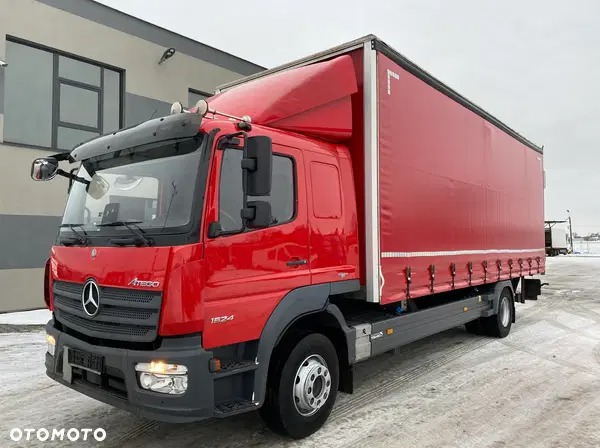 Czerwony samochód ciężarowy Mercedes-Benz Atego 1524 z plandeką, zaparkowany na placu, widoczne detale karoserii i oświetlenia.