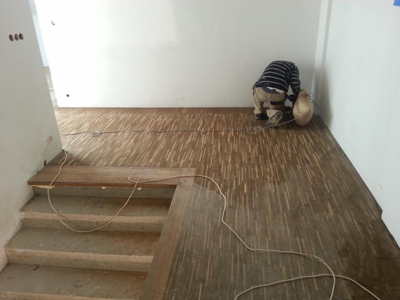 Pracownik szlifuje drewnianą podłogę parkietową w trakcie renowacji, widoczne ślady wilgoci i kabel zasilający na podłodze, schody w tle.