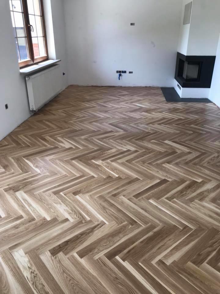 Ukończona podłoga z parkietu ułożonego we wzór jodełki francuskiej w jasnym, nowym wnętrzu z kominkiem i oknem.