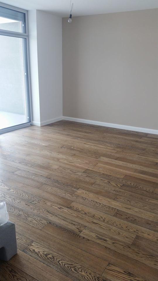 Ułożona podłoga z ciemnego drewna w nowym, jasnym pomieszczeniu z dużym oknem balkonowym, widoczna żarówka na kablu.