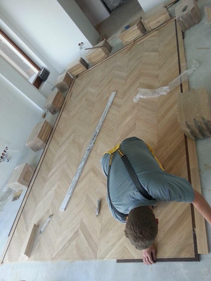 Układanie parkietu w jodełkę; widoczne pakiety desek, listwa pomiarowa i narzędzia, fachowiec przy pracy.