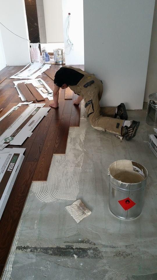 Montaż ciemnych desek podłogowych na klej, widoczny pracownik w kombinezonie, puszki z klejem i narzędzia.