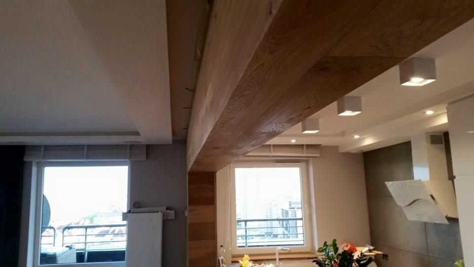 Wnętrze mieszkania z drewnianą belką stropową i drewnianą okładziną ściany, widok na okno z widokiem na miasto.