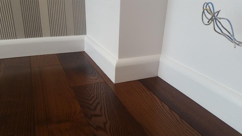 Ciemne drewniane deski podłogowe z białymi listwami przypodłogowymi, widoczne wystające przewody elektryczne na ścianie w tle.