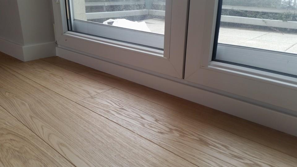 Jasne drewniane deski podłogowe ułożone równolegle do białego, przesuwanego okna balkonowego z widokiem na taras.