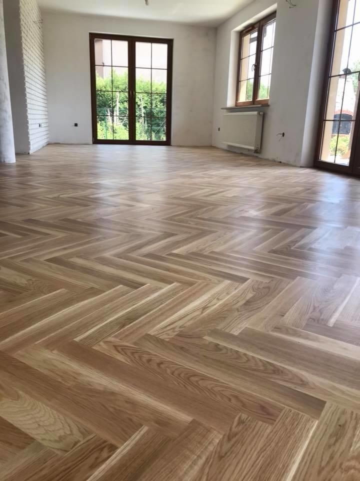 Jasna, parkietowa podłoga ułożona we wzór jodełki francuskiej w przestronnym, częściowo wykończonym pokoju z dużymi oknami.