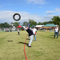 Mężczyzna w trakcie rzutu oponą na trawiastym terenie podczas imprezy plenerowej z dmuchańcem, namiotami i uczestnikami w tle.