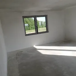 Surowe wnętrze pokoju z oknem, ściany pokryte szarym tynkiem, widoczne ślady po gniazdkach elektrycznych, betonowa podłoga z cieniem od okna.