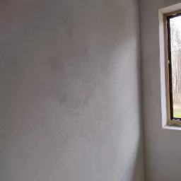 Szara, surowa ściana w rogu pokoju z widoczną strukturą, oświetlona naturalnym światłem wpadającym przez okno z drewnianą ramą, ukazująca minimalizm wnętrza.