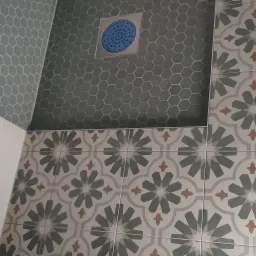 Wnętrze łazienki z mozaiką heksagonalną w odcieniach szarości i niebieskim odpływem liniowym oraz dekoracyjnymi płytkami z motywem kwiatowym w kolorach szarości, beżu i bieli.