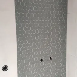 Ściana łazienki wyłożona szarymi, heksagonalnymi płytkami mozaikowymi z widocznymi otworami na instalacje wodno-kanalizacyjne, sąsiadująca z białą, gładką ścianą.