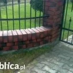 Ceglany słupek z czarnym, metalowym ogrodzeniem na tle zielonego trawnika i kostki brukowej. Słupek wykończony z czerwonej cegły klinkierowej.