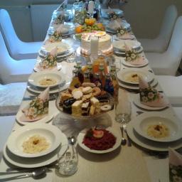 KONKRET B&M CATERING - Uroczysty stół z białym obrusem, nakryty dla wielu gości, z ciastami na paterze, napojami w butelkach, talerzami z daniami i tortem z zapaloną świeczką, otoczony białymi krzesłami.