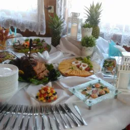 Stół z białym obrusem, zastawiony różnorodnymi potrawami, w tym pieczonym mięsem, serami, owocami, przekąskami na paterach, dekoracjami w postaci ananasów i białych lampionów, oraz ułożonymi...