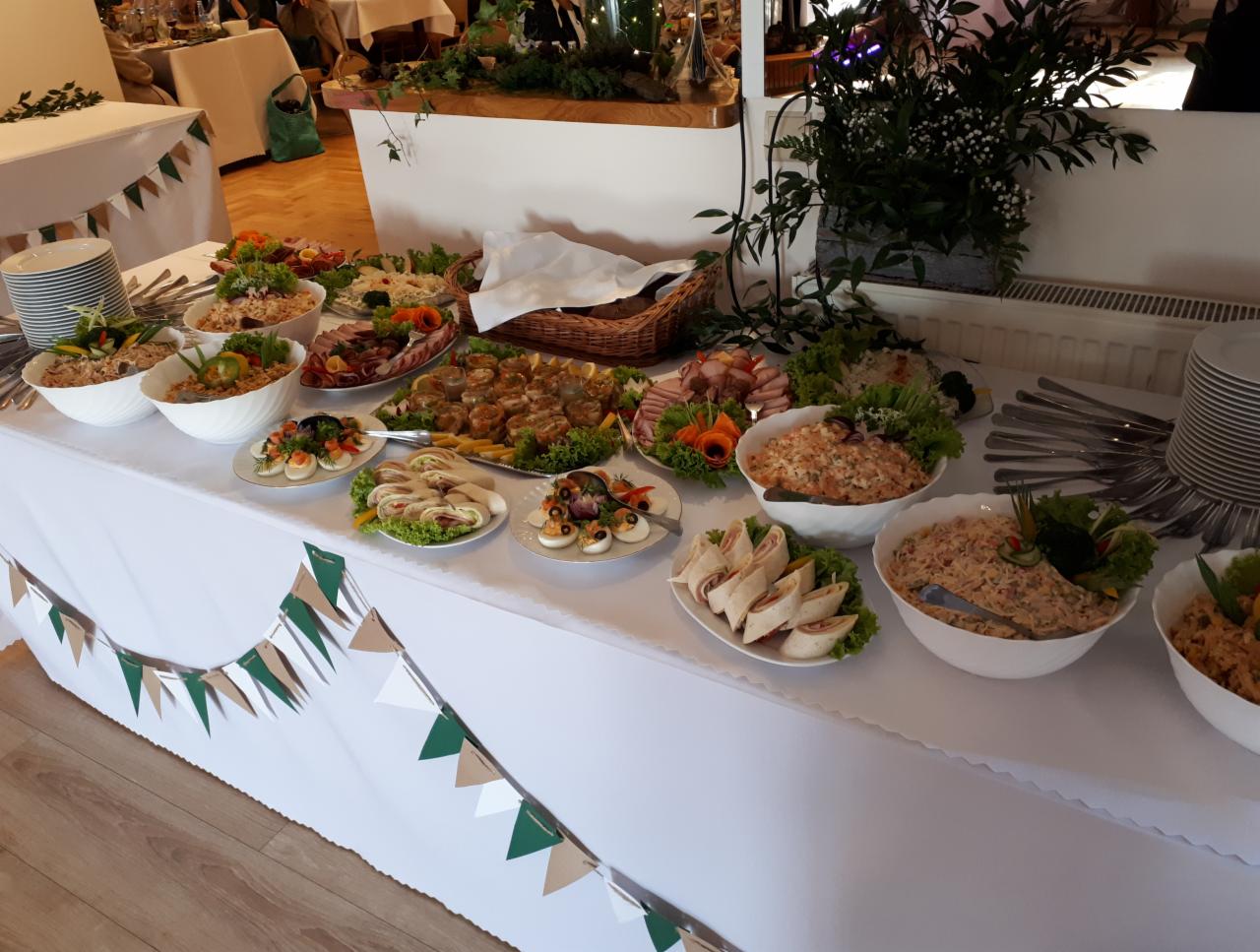 Stół bufetowy z różnorodnymi przekąskami, sałatkami i daniami na przyjęciu, udekorowany girlandą z papierowych chorągiewek w kolorach zieleni i beżu.