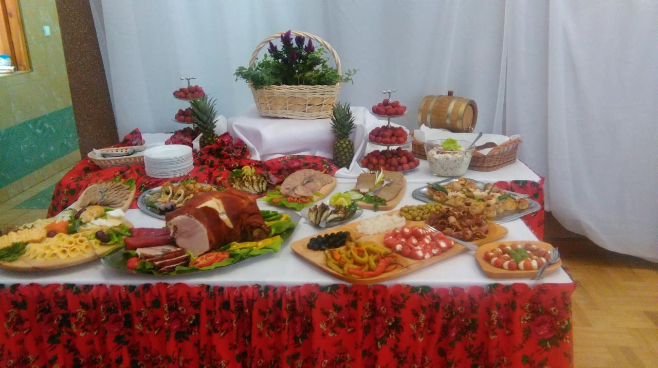 Stół cateringowy z wędlinami, serami, sałatkami i owocami, dekorowany kwiatami w koszyku i ananasami, z czerwonym obrusem w kwiaty.