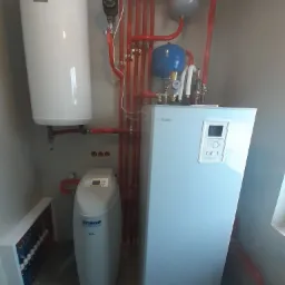 Instalacja grzewcza w kotłowni z białym zasobnikiem ciepłej wody, szarym zbiornikiem wyrównawczym i niebieskim naczyniem przeponowym, połączonymi czerwonymi rurami, widoczny również zmiękczacz wody.