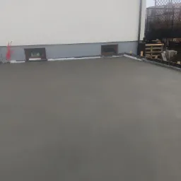 Świeżo wylana, gładka posadzka jastrychowa przed budynkiem z widocznymi przewodami elektrycznymi i oknami piwnicznymi.