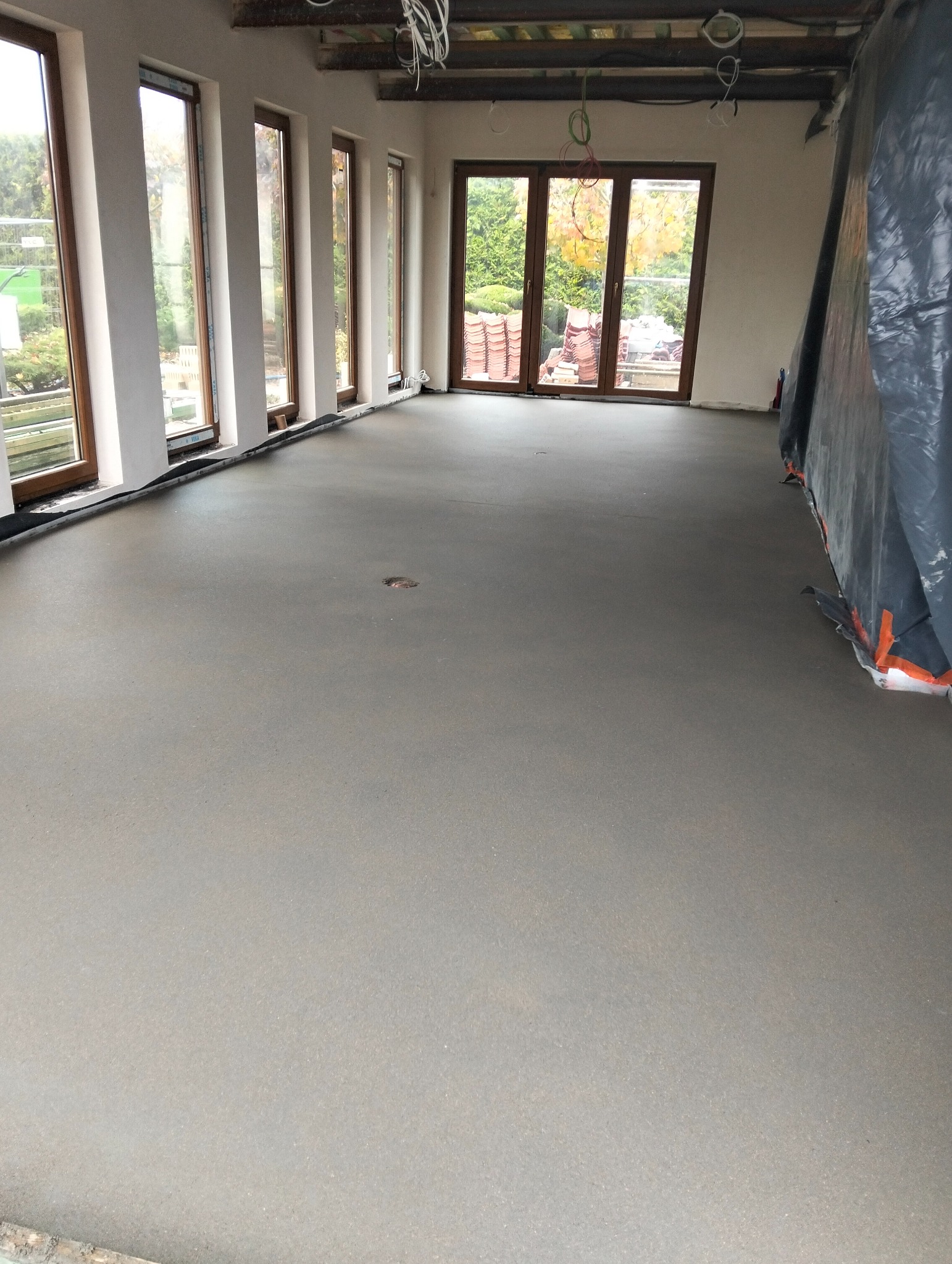 Świeżo wylana, gładka, szara posadzka w jasnym pomieszczeniu z dużymi oknami i widokiem na drzewa; zabezpieczona folią część podłogi po prawej.