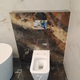 Nowoczesna, podwieszana toaleta w łazience wykończonej marmurowymi płytami w odcieniach bieli, brązu i czerni, z widocznym przyciskiem spłukującym w obudowie z płytek oraz częścią umywalki po lewej...