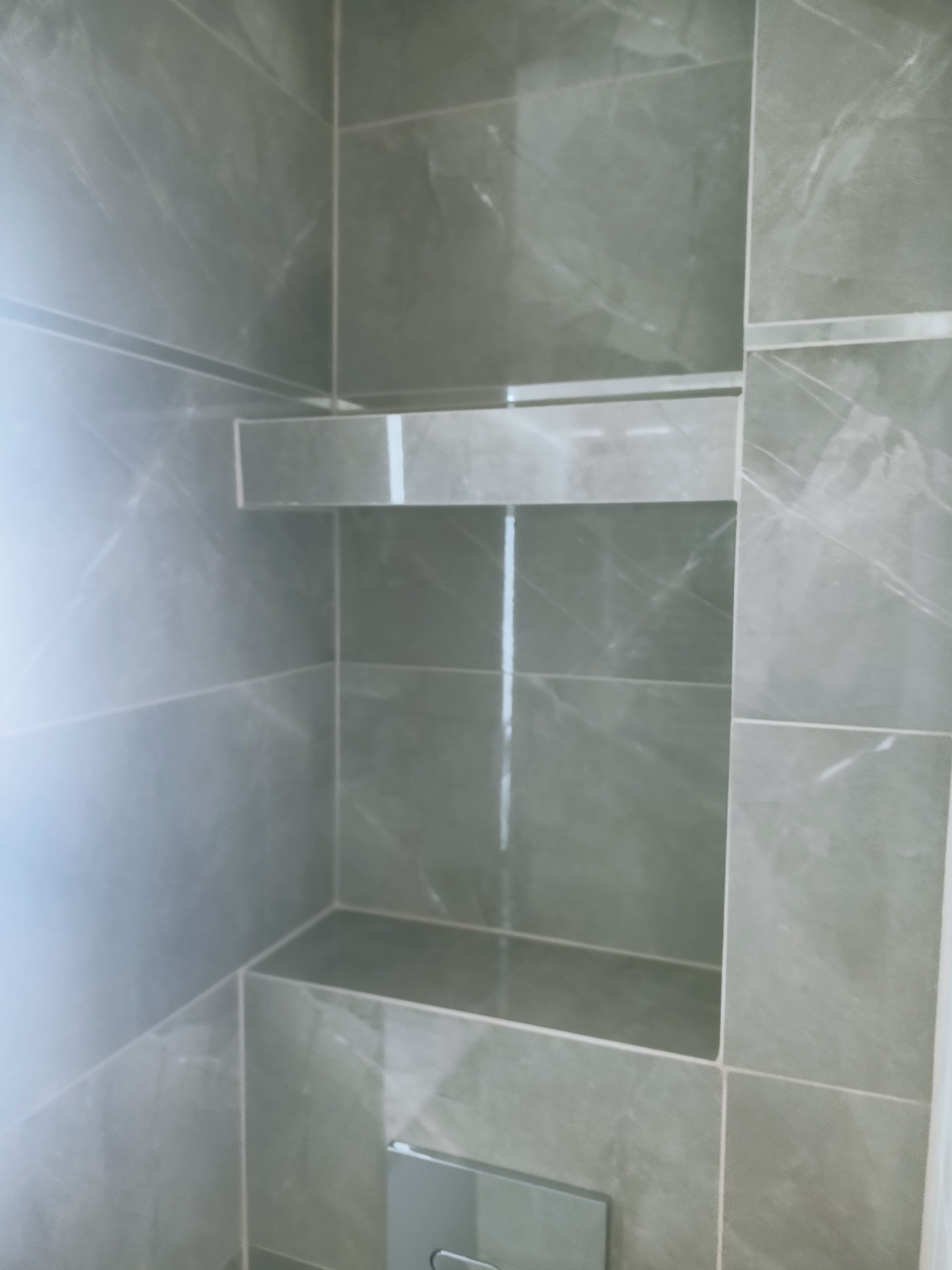 Szare, marmurkowe płytki w łazience z wnęką wyłożoną tymi samymi płytkami i półką.