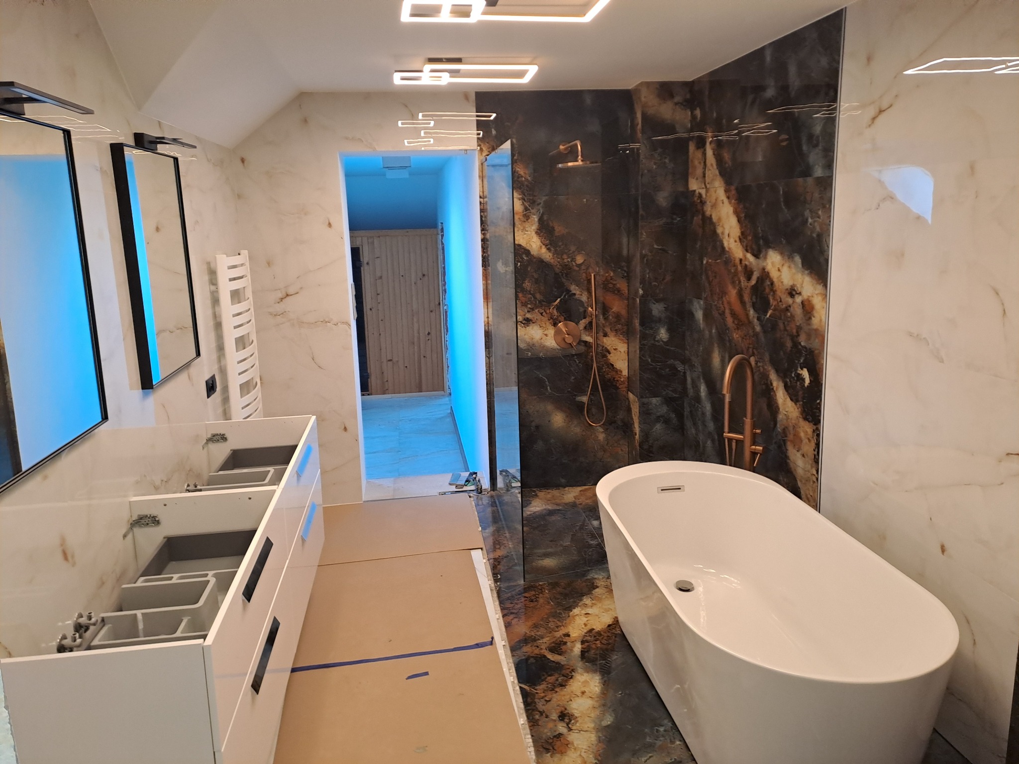 Nowoczesna łazienka w trakcie remontu: biała wanna wolnostojąca, marmurowe płytki ścienne w odcieniach beżu i brązu, podwieszana szafka umywalkowa z otwartymi szufladami, oświetlenie LED w suficie,...