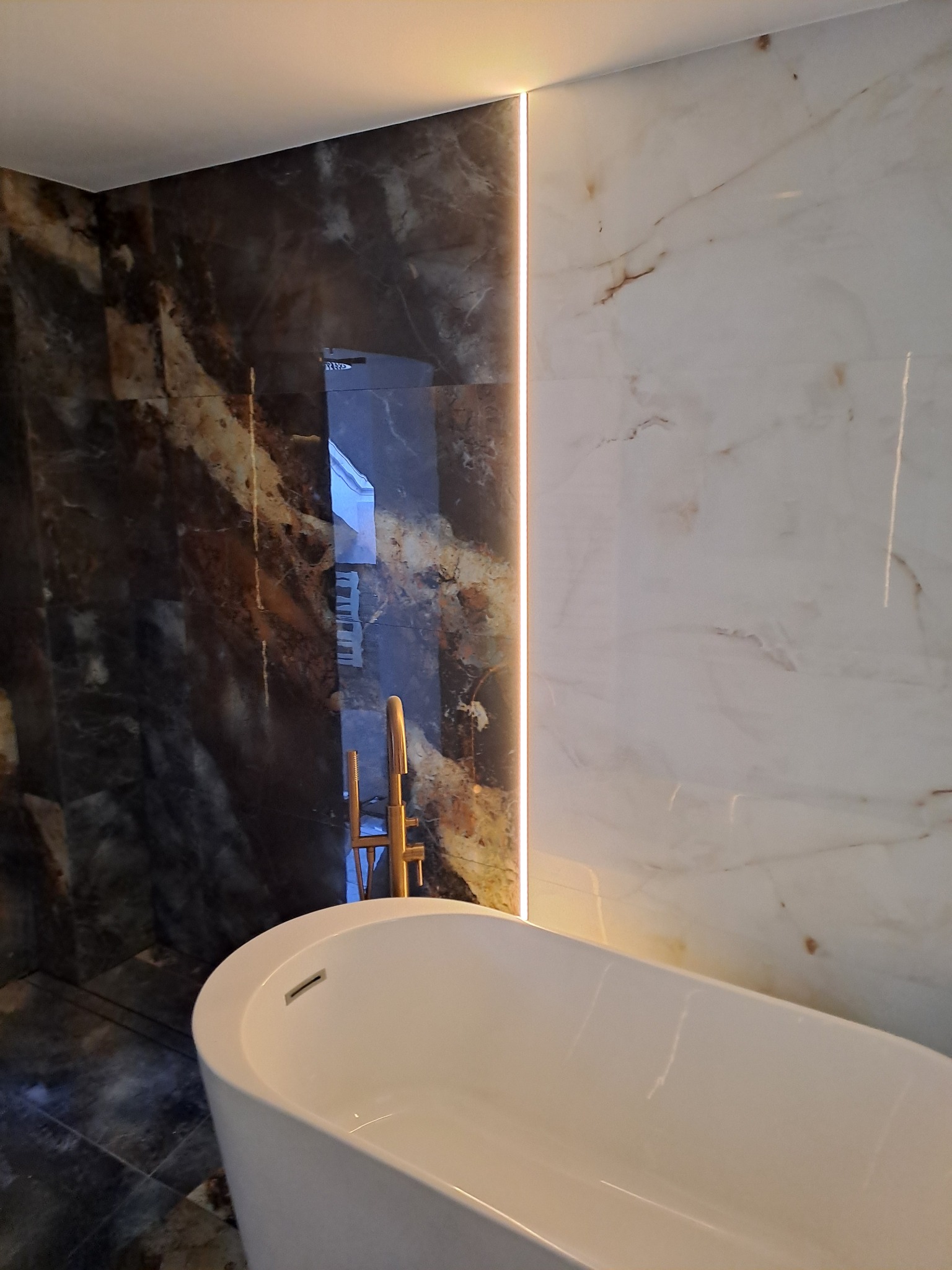 Nowoczesna łazienka z wolnostojącą białą wanną, marmurowymi płytkami w odcieniach brązu i bieli, podświetleniem LED w pionowej szczelinie między płytkami oraz złotą armaturą widoczną w odbiciu lustra.