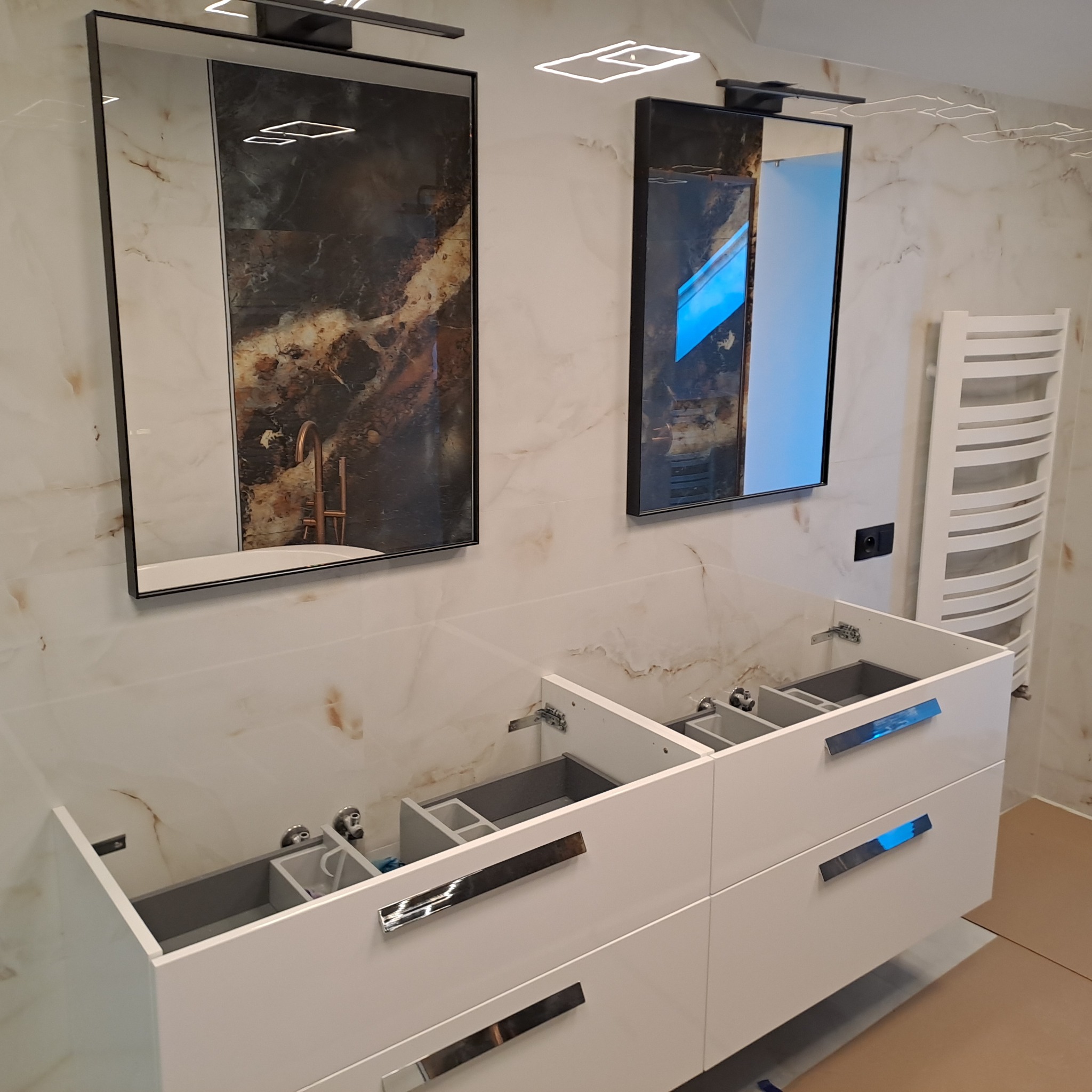 Łazienka w trakcie montażu: biała szafka pod umywalkę z chromowanymi uchwytami, dwa lustra w czarnych ramach z oświetleniem, marmurowe płytki na ścianach i grzejnik drabinkowy.