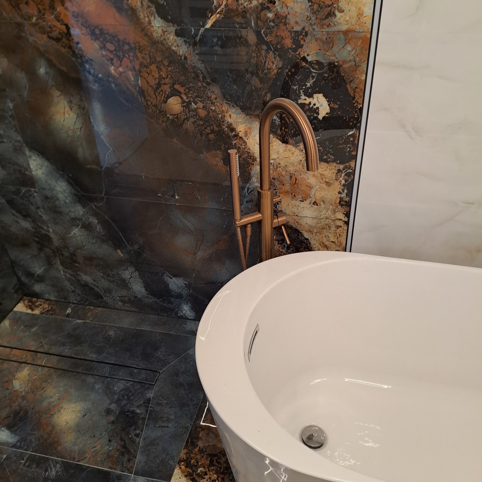 Wolnostojąca wanna z brązową armaturą na tle ściany z płytek imitujących marmur w odcieniach brązu, czerni i bieli.