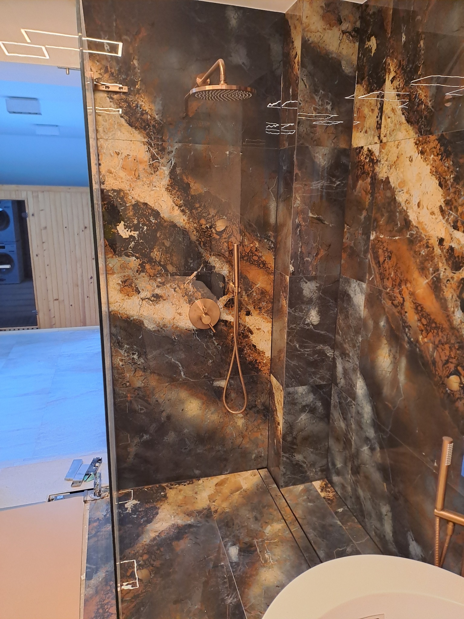 Kabina prysznicowa z marmurowymi płytkami w odcieniach brązu i czerni, z miedzianą armaturą, widok z zewnątrz kabiny na fragment łazienki z drewnianą boazerią i pralką.