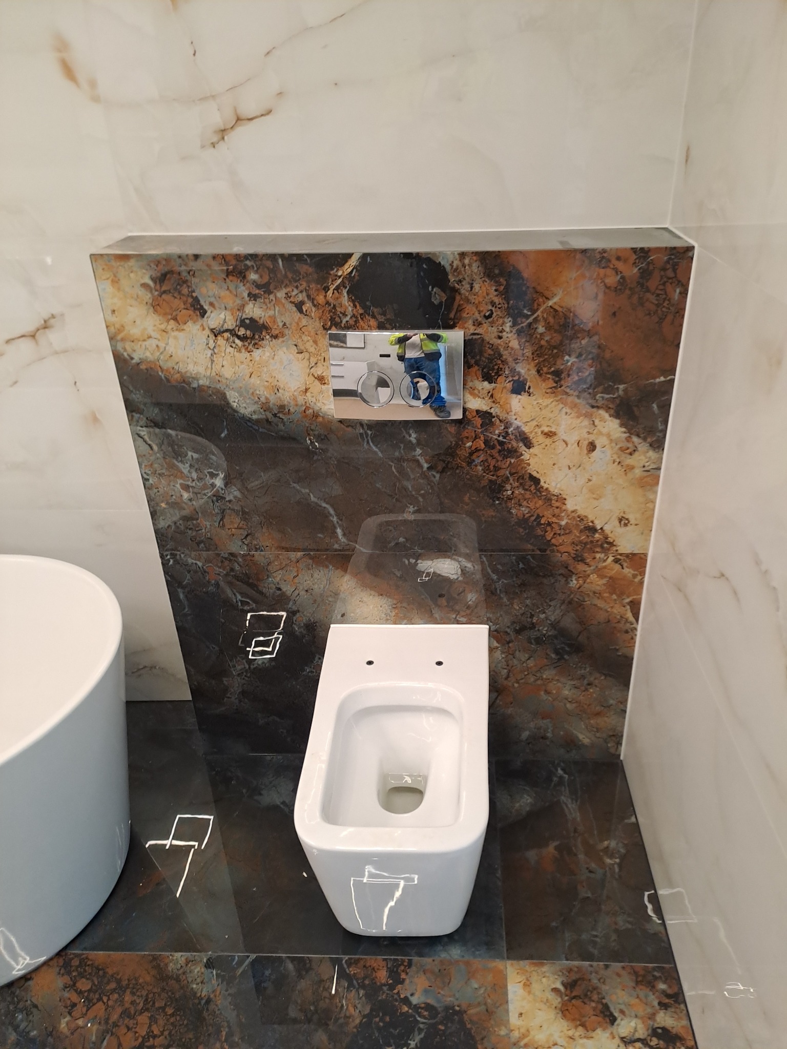 Nowoczesna, podwieszana toaleta w łazience wykończonej marmurowymi płytami w odcieniach bieli, brązu i czerni, z widocznym przyciskiem spłukującym w obudowie z płytek oraz częścią umywalki po lewej...