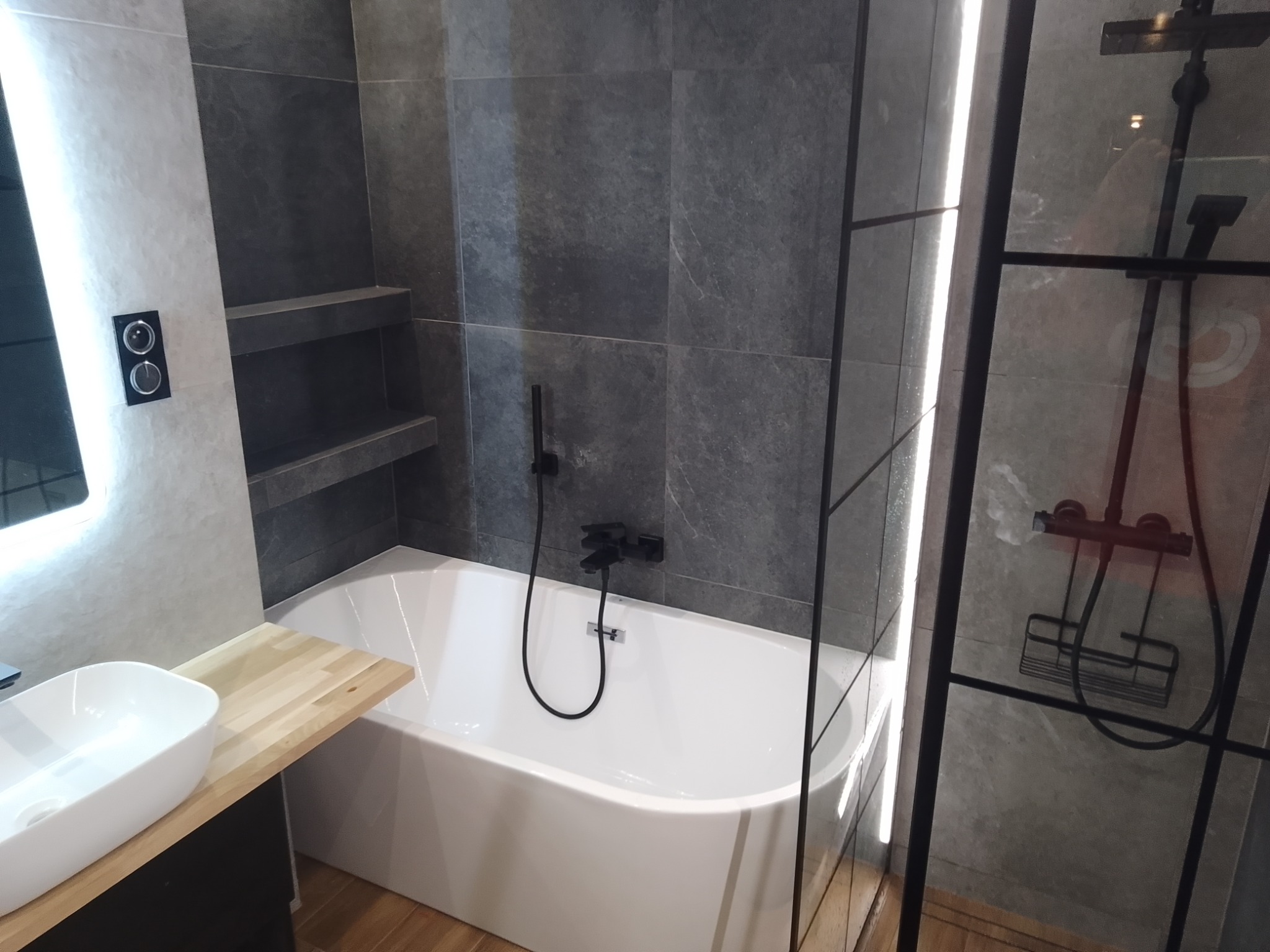 Nowoczesna łazienka z wanną, czarną armaturą i szarymi płytkami imitującymi kamień, półki wnękowe w ścianie, umywalka nablatowa na drewnianym blacie.