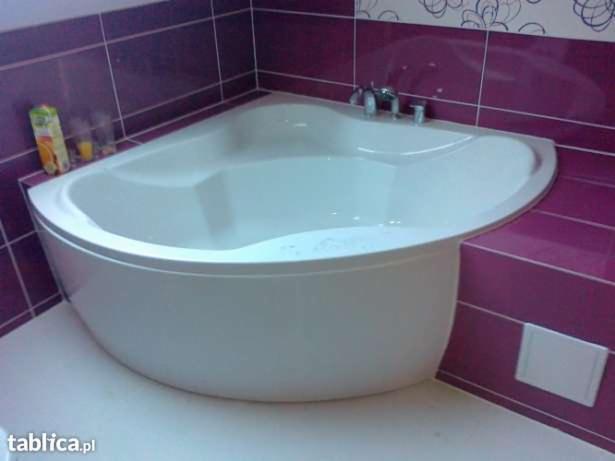 Narożna wanna akrylowa z chromowaną armaturą, obudowana i otoczona fioletowymi płytkami ceramicznymi z dekoracyjnym wzorem na górze, widoczne kosmetyki i akcesoria łazienkowe.