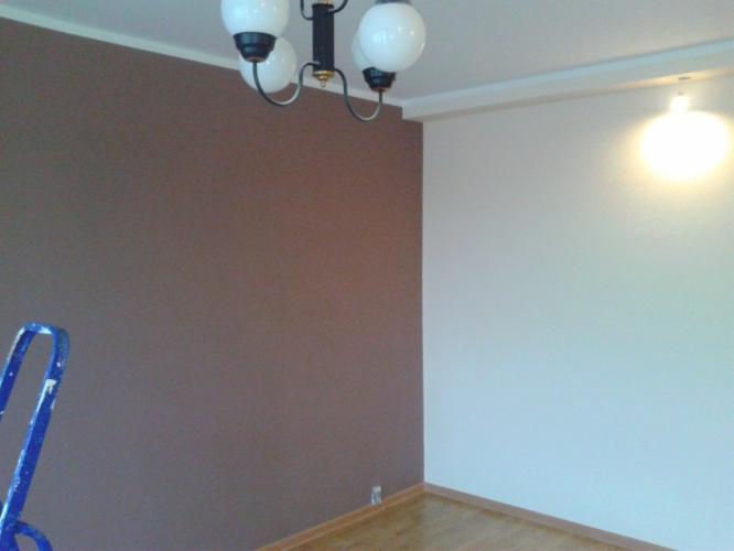 Pokój po częściowym pomalowaniu ścian: jedna ściana w kolorze brązowym, druga biała, widoczna drabina malarska i lampa sufitowa.