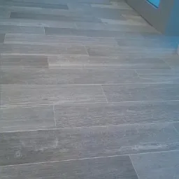 Ułożona szara podłoga z płytek imitujących drewno, widok z góry. Fragment szafki i drzwi w tle. Płytki ułożone w regularny wzór.