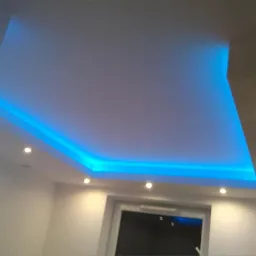 Nowoczesny sufit podwieszany z niebieskim oświetleniem LED, widoczne punktowe lampy halogenowe, ściany pomalowane na biało, okno w tle.