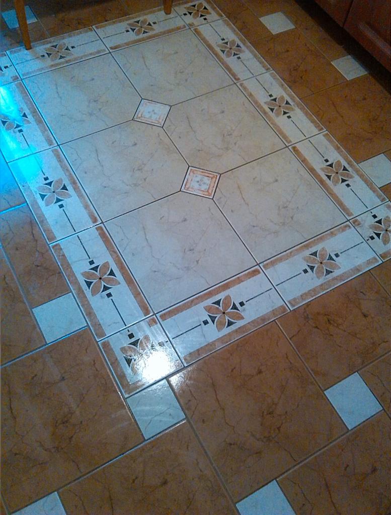 Ułożona podłoga z płytek w dwóch odcieniach brązu, z dekoracyjnym wzorem kwiatowym na kremowej części pośrodku. Widok z góry, fragment krzesła w tle.