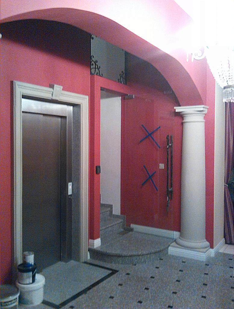 Wnętrze holu z kolumną i windą w budynku. Ściany w odcieniach różu, podłoga z lastryko. Widoczne schody i fragment żyrandola. Drzwi zabezpieczone taśmą.