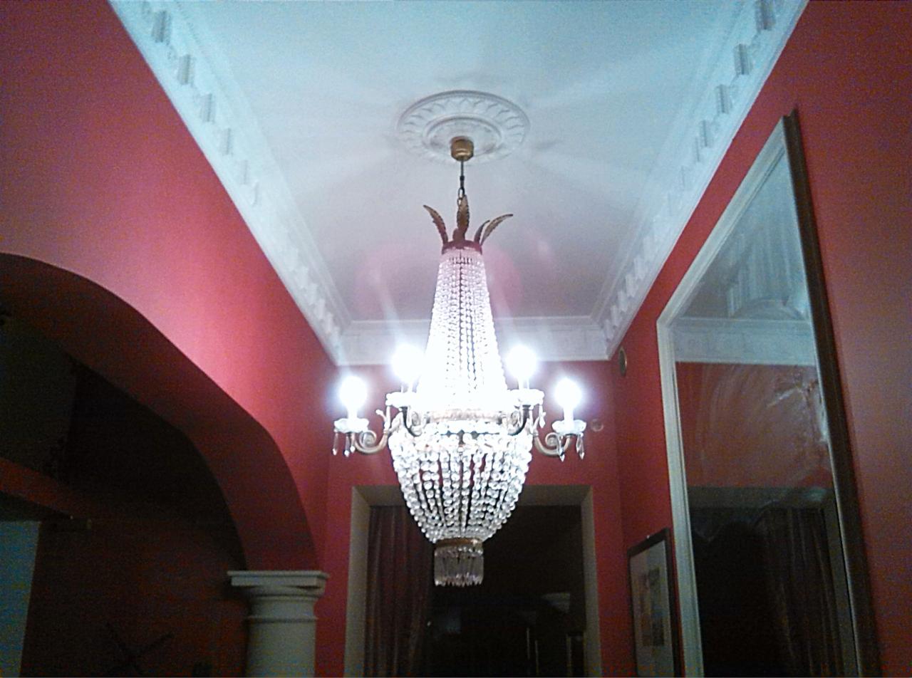 Kryształowy żyrandol w holu z dekoracyjnym sufitem i czerwonymi ścianami. Widoczny fragment kolumny i rama lustra. Ujęcie z dołu.