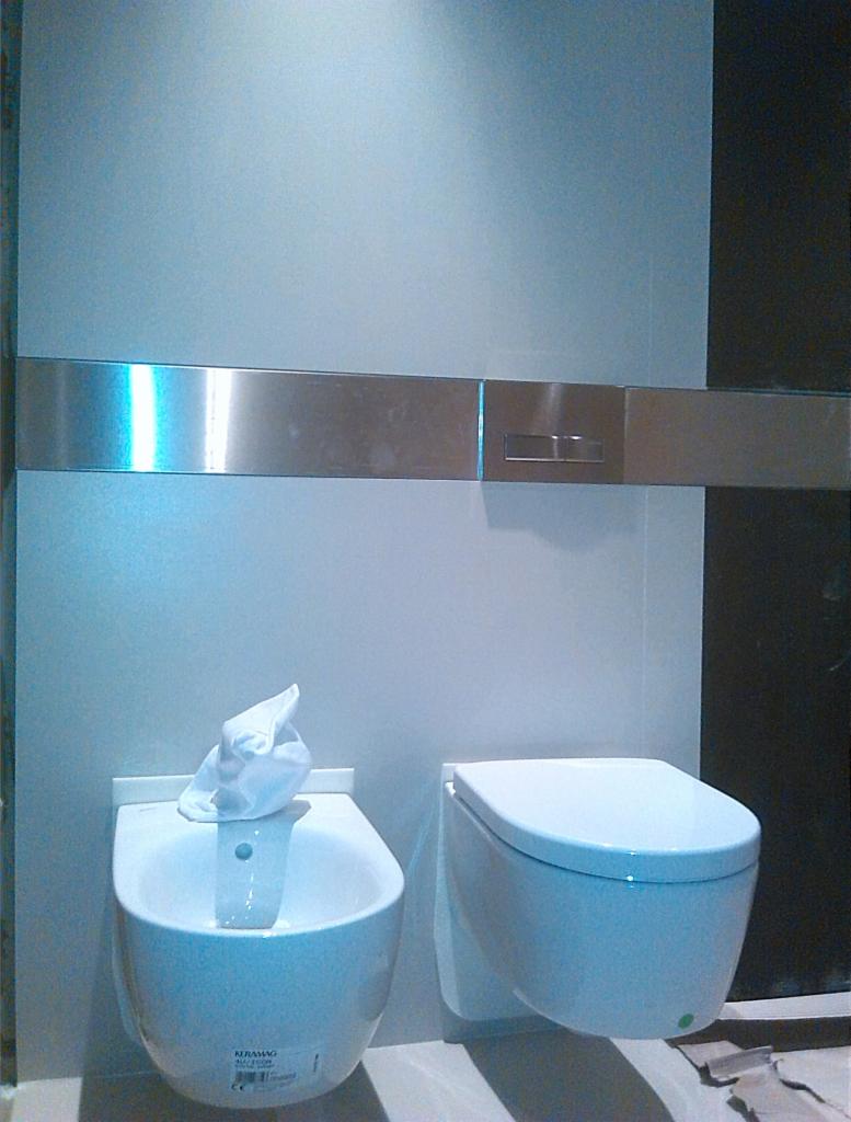 Nowoczesna łazienka: białe bidet i toaleta podwieszane, chromowana listwa z przyciskami spłukiwania, minimalistyczny design, chłodne oświetlenie.